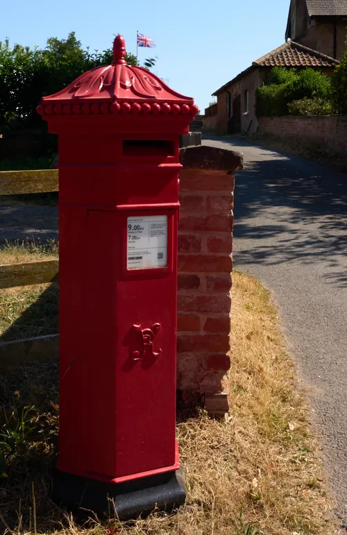 This architectural photograph captures a traditional British red post box situated on the edge of a quiet village lane. The post box is positioned next to a brick wall, with a wooden fence visible on the left, and houses with tiled roofs lining the background. Above the post box, a British flag is prominently flying, reinforcing the setting’s national identity. The bright sunlight and clear blue sky, along with the dry grass, indicate that the picture was taken during the afternoon in midsummer. The overall scene showcases quintessential rural England.
